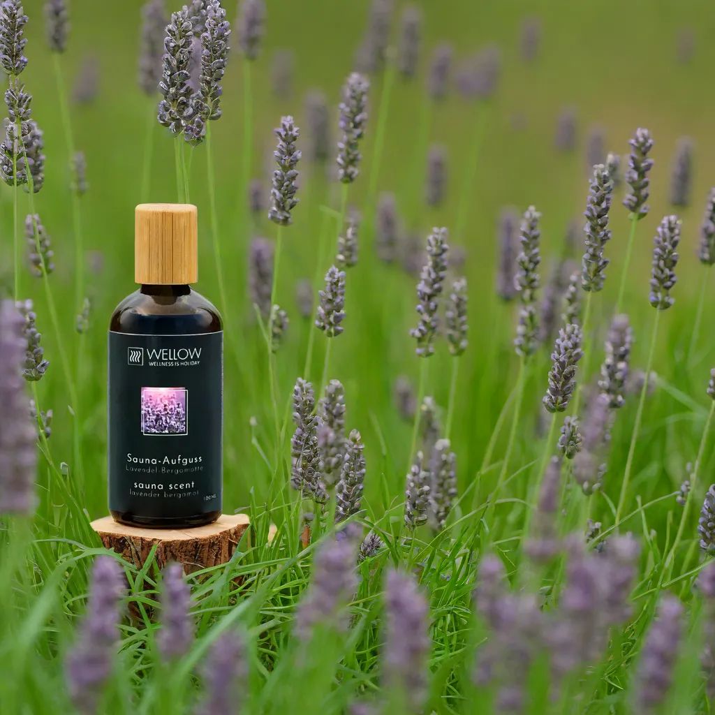 Flacone di vetro marrone con tappo in legno su ceppo di legno. Etichetta nera con testo e logo. Fiori di lavanda sullo sfondo.
