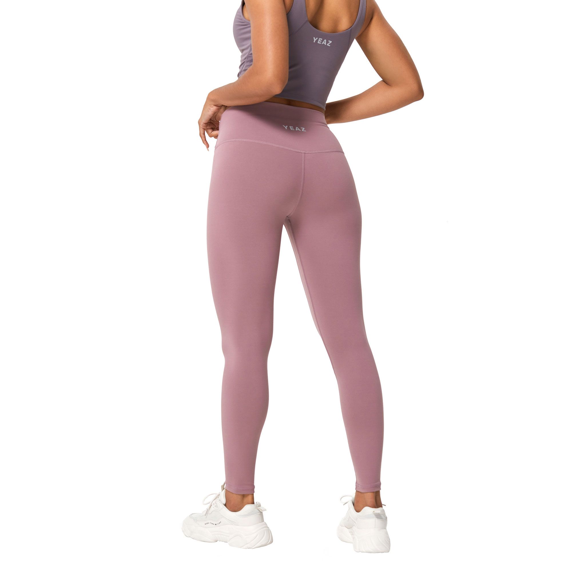 Una donna indossa un leggings rosa e un top coordinato con il logo 'YEAZ'. Scarpe da ginnastica bianche completano l'outfit.