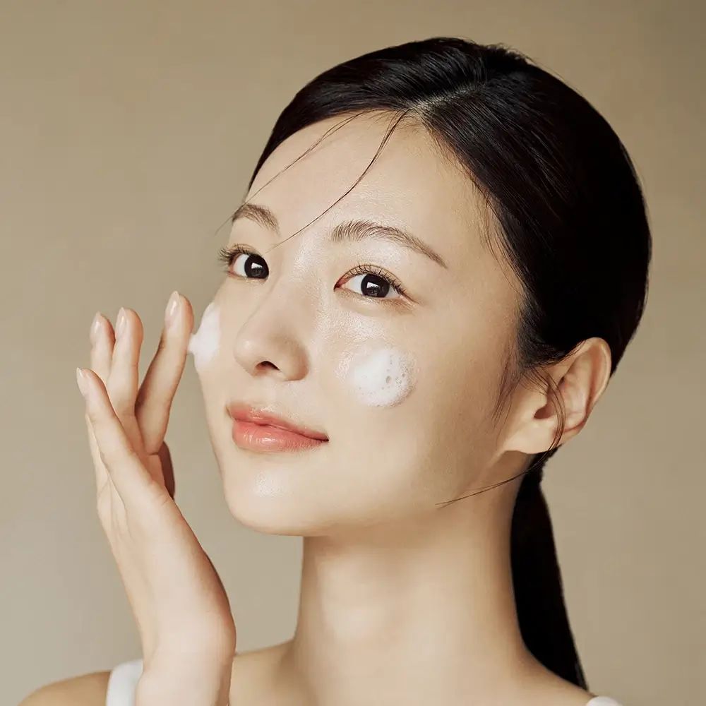 Una donna applica schiuma sul viso. Ha i capelli scuri e una canotta bianca.