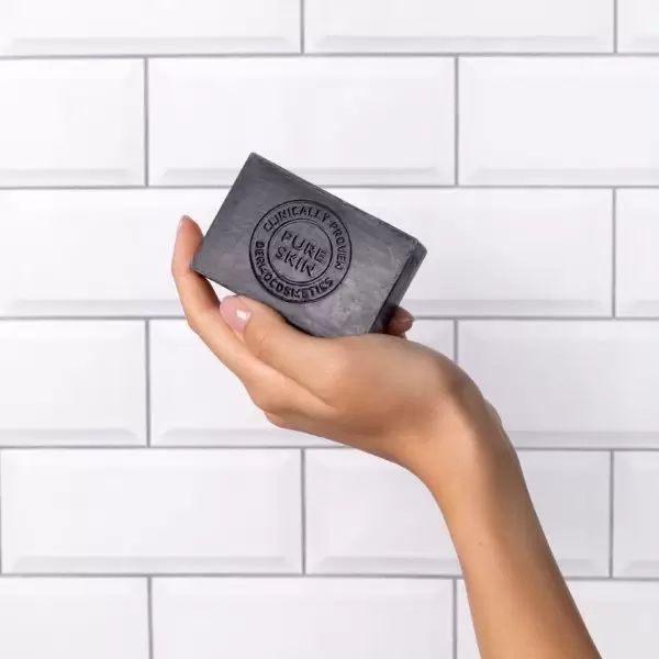 Sapone nero in mano. Scritta: Pure Skin, clinicamente testato. Forma rettangolare. Sfondo: piastrelle bianche.