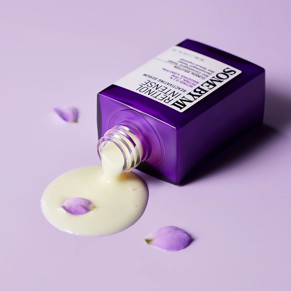 Flacone viola sdraiato, siero che fuoriesce. Scritta: Retinol Intense Reactivating Serum. Marchio: SOMEBYMI. Petali viola.