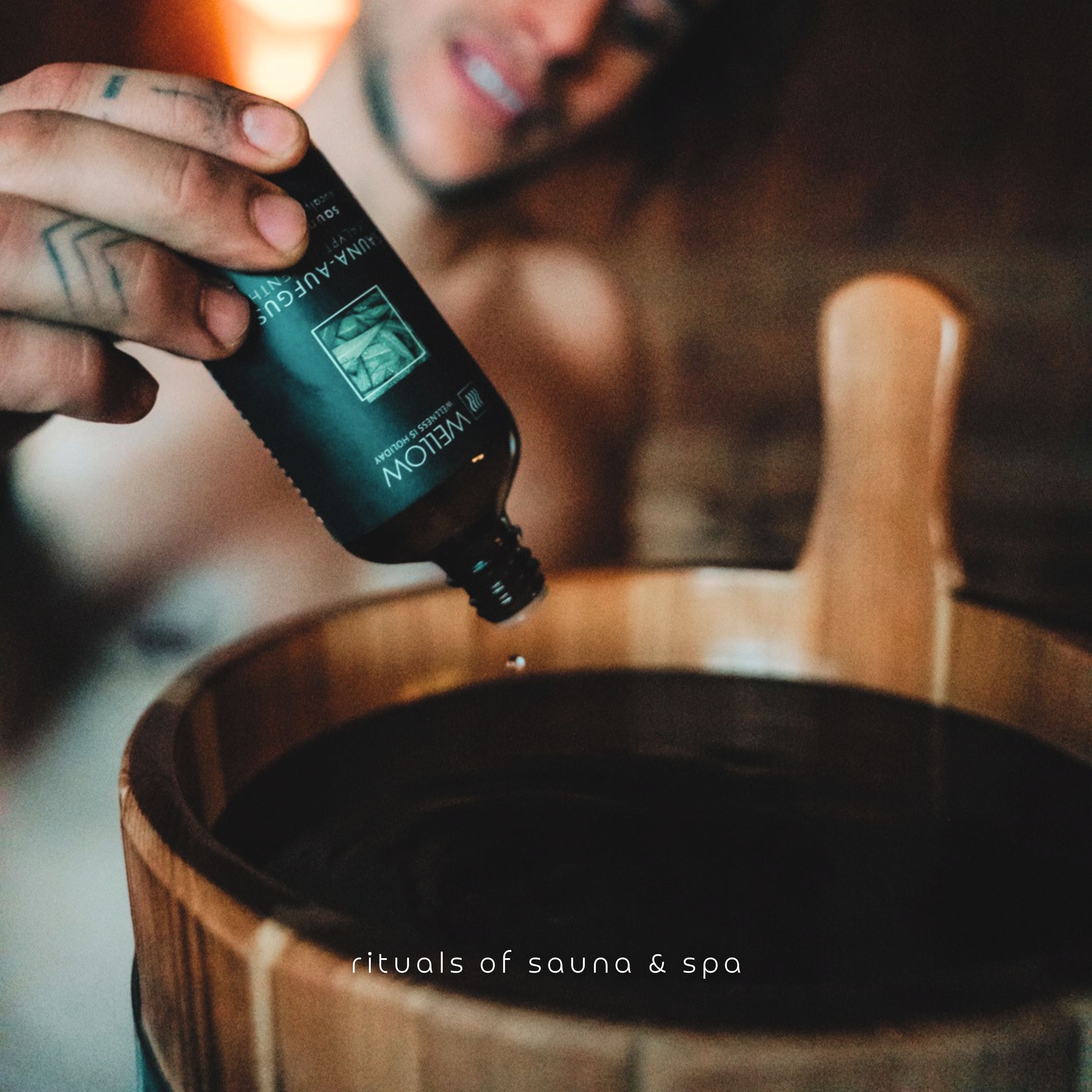 Un uomo versa del liquido da una bottiglia in un secchio di legno. Scritta sulla bottiglia: Sauna-Aufguss.