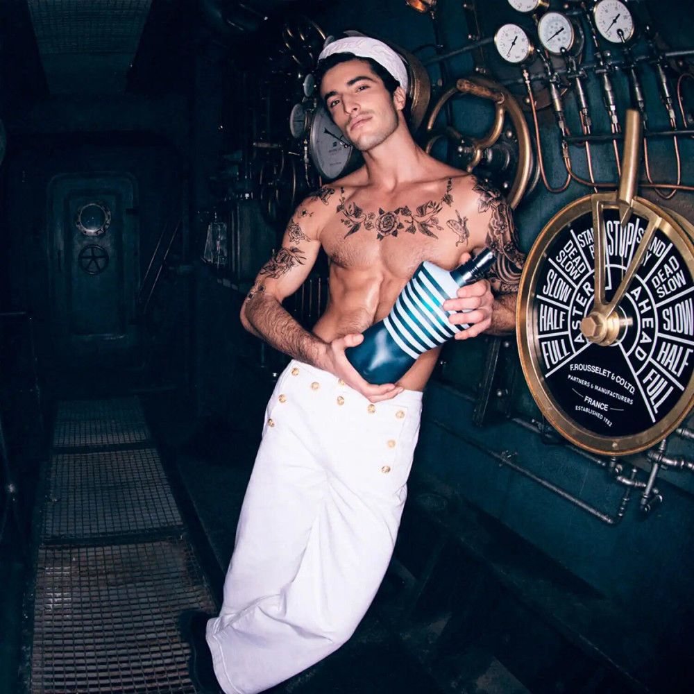 Uomo tatuato in abito da marinaio tiene in mano un flacone. In background: timone e strumenti.