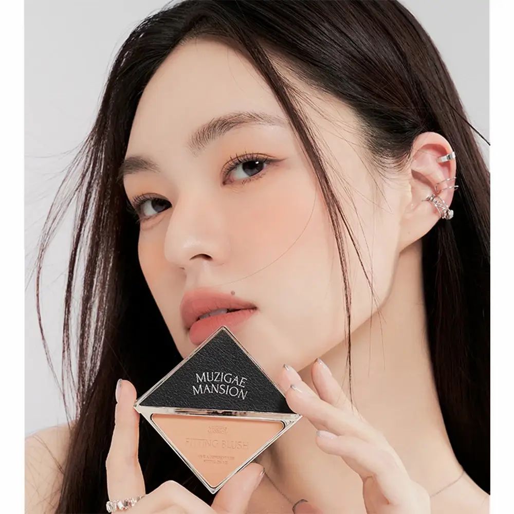 Persona tiene MUZIGAE MANSION Fitting Blush. Confezione triangolare nera e argento. Fard in tonalità pesca visibile.