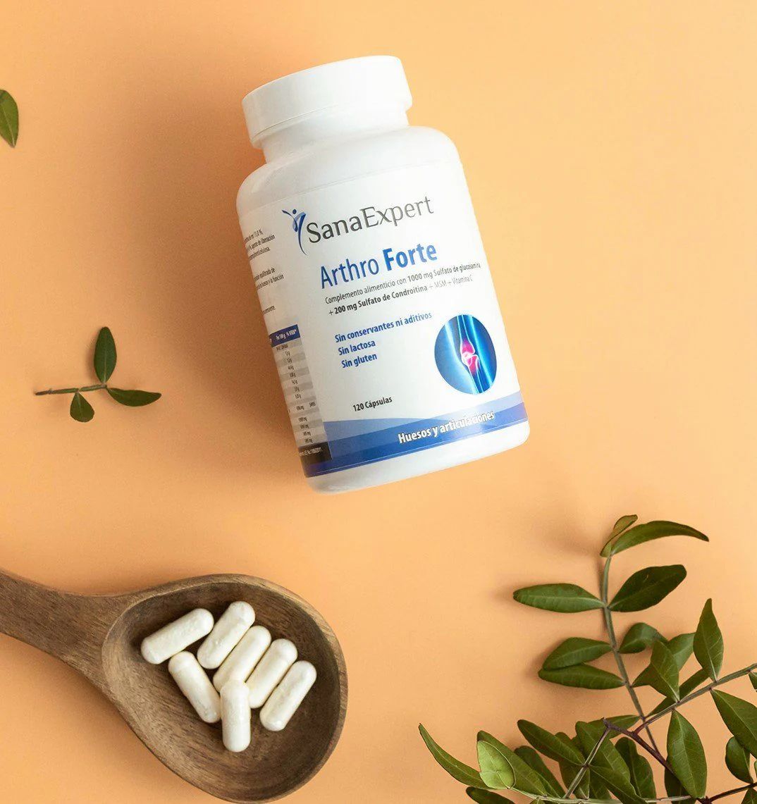 Flacone bianco SanaExpert Arthro Forte con capsule su un cucchiaio di legno. Piante verdi decorano l'immagine.