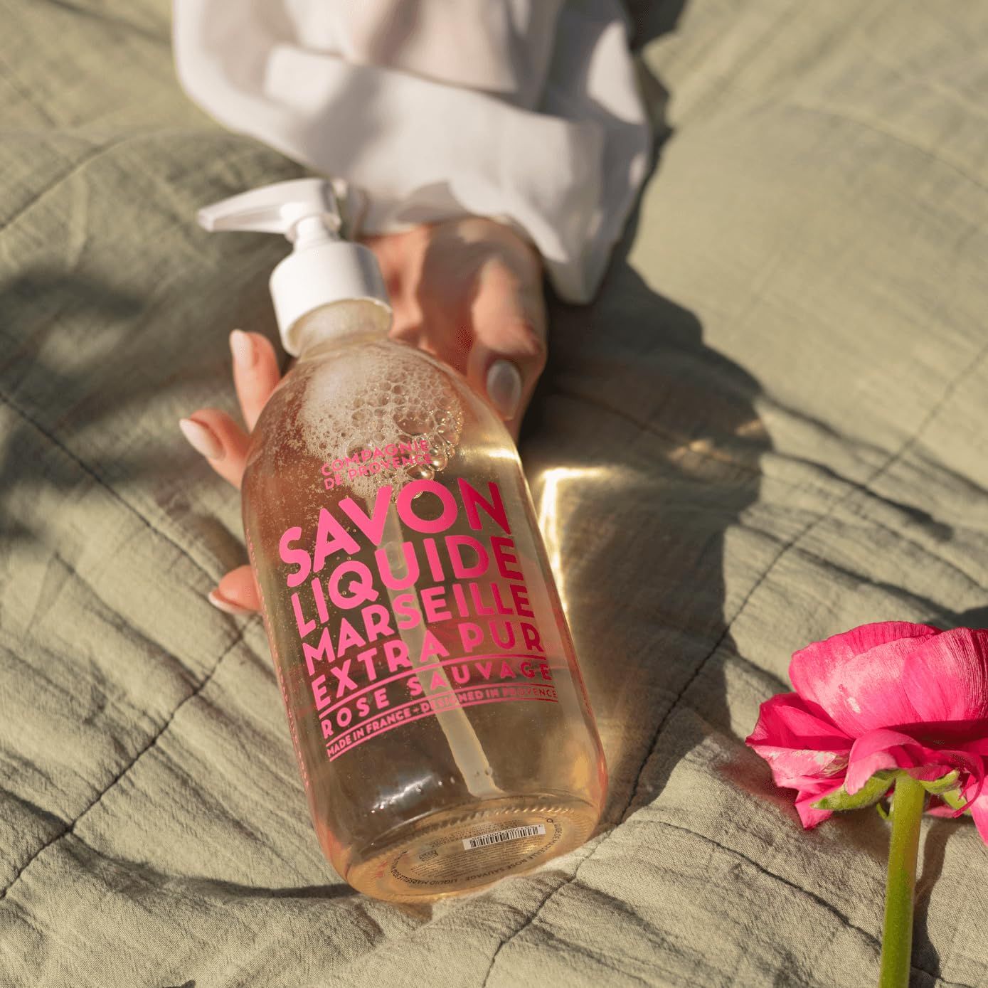 Una mano tiene un flacone trasparente con etichetta rosa. Scritta: Savon Liquide Marseille Extra Pur Rose Sauvage. Accanto, una rosa.
