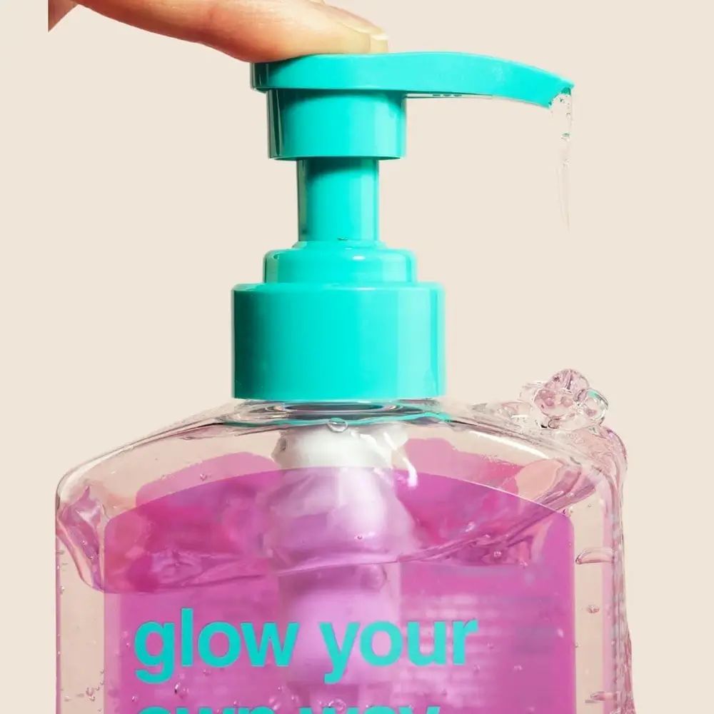 Primo piano di un flacone trasparente con liquido rosa. Pompa blu. Liquido che gocciola. Scritta: glow your own way.