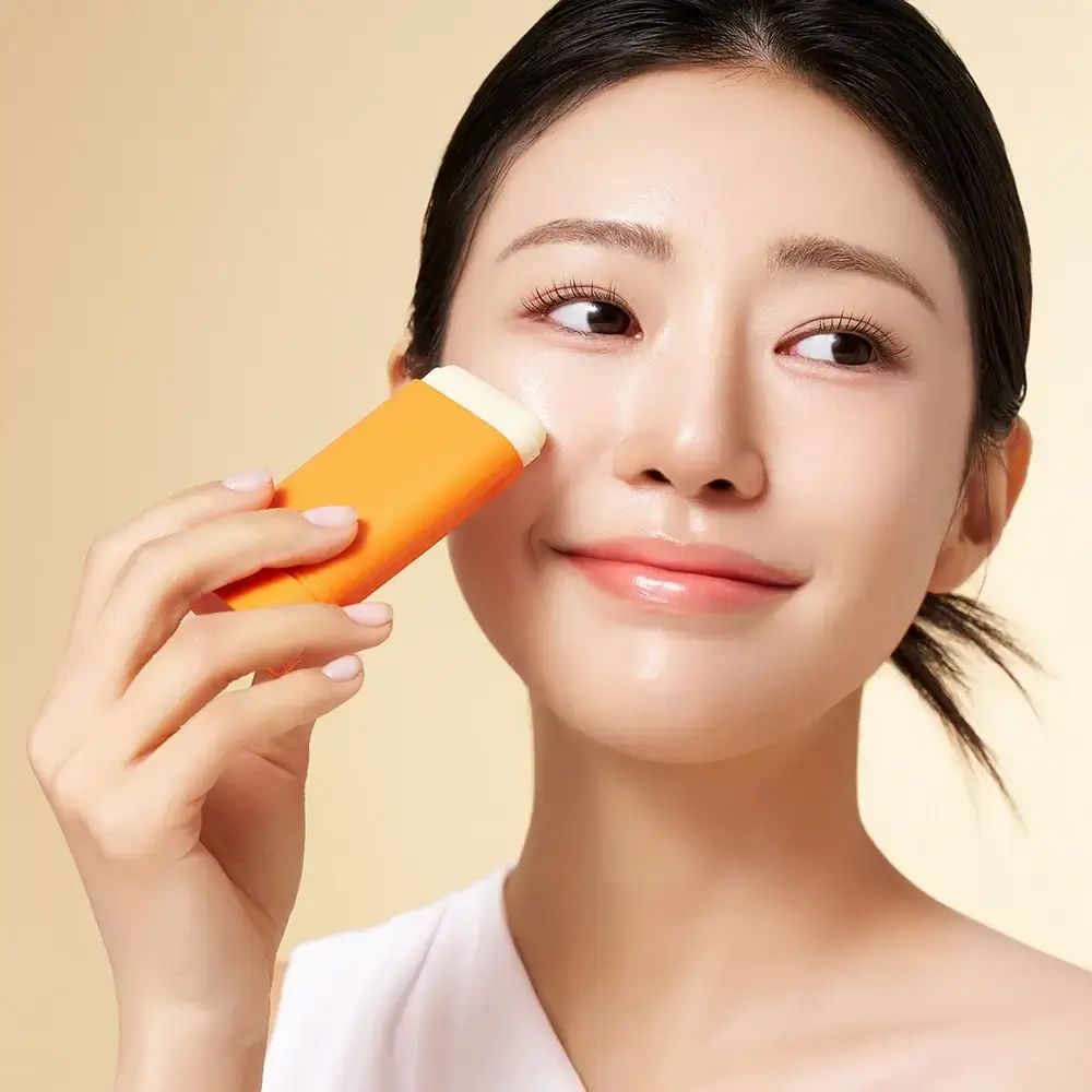 Donna applica stick arancione sulla guancia. Lo stick ha un'estremità bianca.