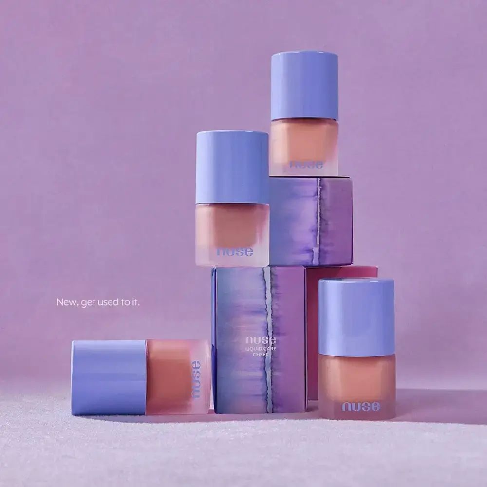 Diversi flaconi e confezioni di NUSE Liquid Care Cheek. Flaconi con tappo blu e contenuto rosa. Confezioni viola.