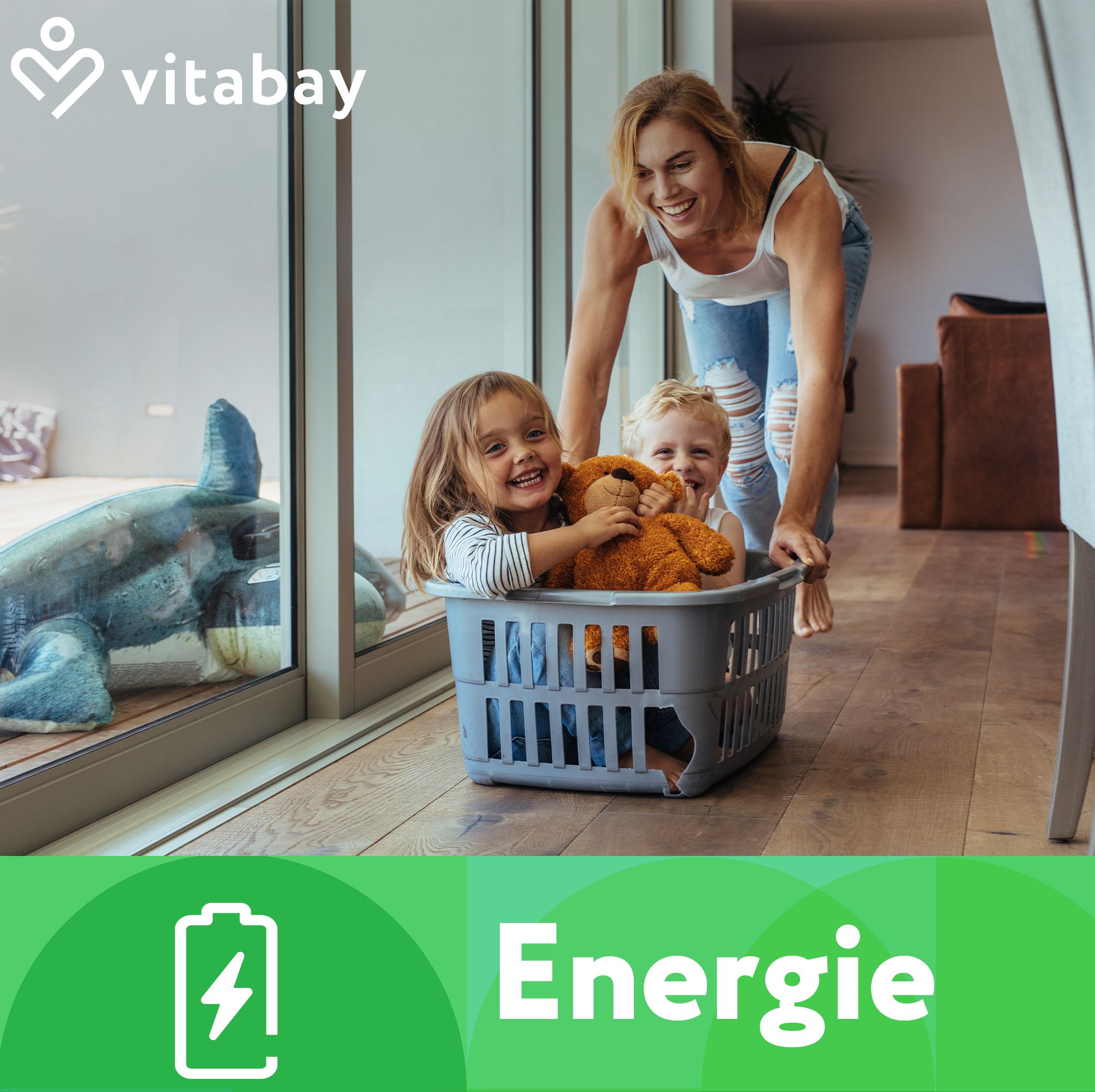 Logo Vitabay. Testo: Energia. Donna spinge bambini in un cesto della biancheria. Bambini che ridono. Sfondo: finestra, soggiorno.