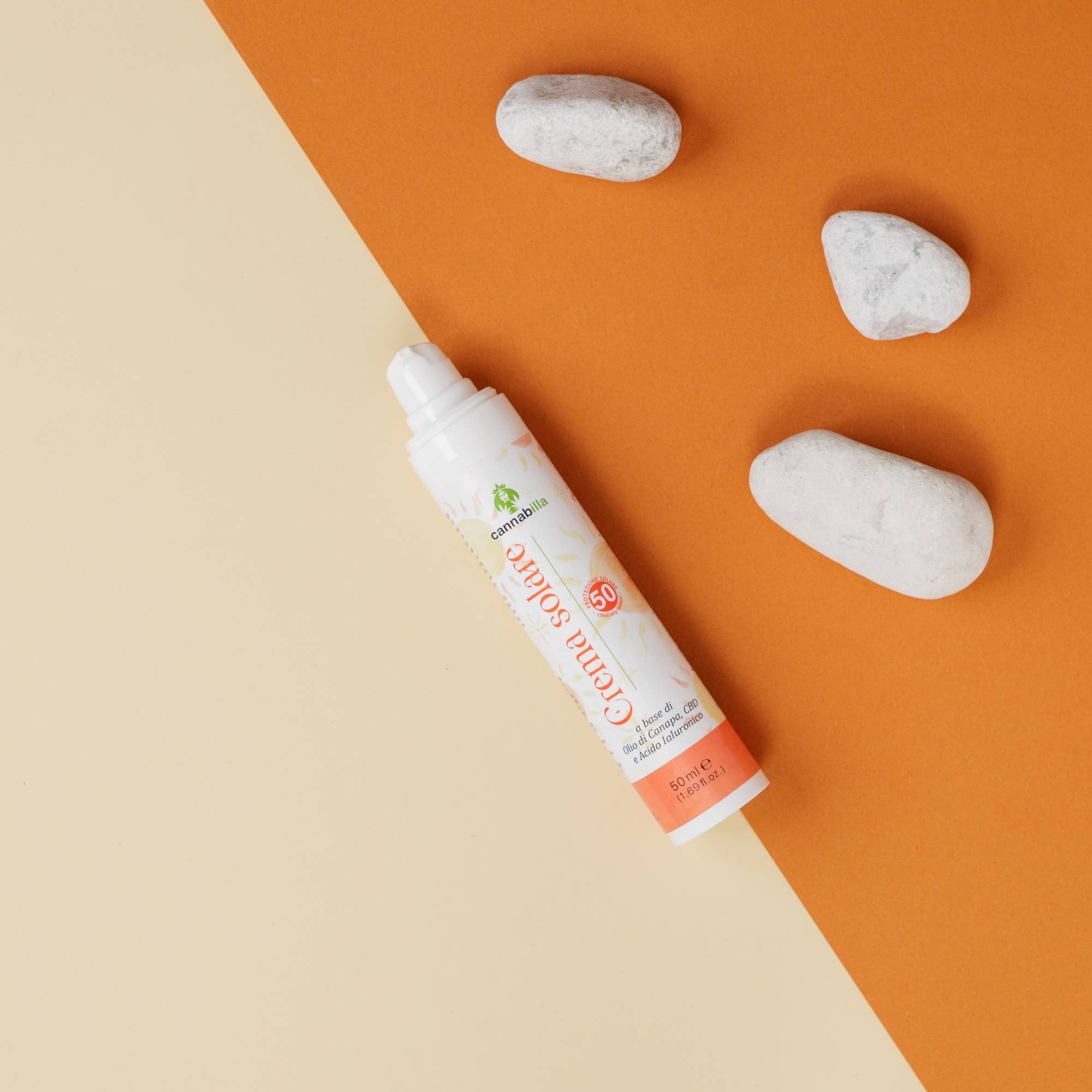 Flacone bianco con dosatore su sfondo bicolore. Scritta: Crema solare, SPF50. Marchio: Cannabilla. Con pietre.