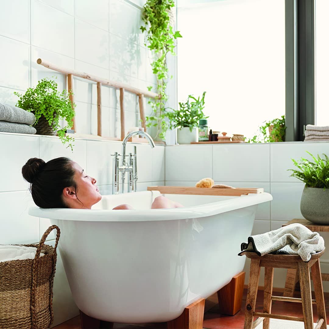 Donna in vasca da bagno bianca con scala in legno e piante. Vasca su gambe in legno. Asciugamani grigi e sgabello in legno.