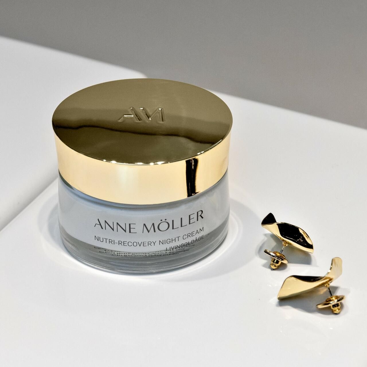 Vaso di crema con coperchio dorato e orecchini. Scritta: Anne Möller, Nutri-Recovery Night Cream, Livingoldâge.