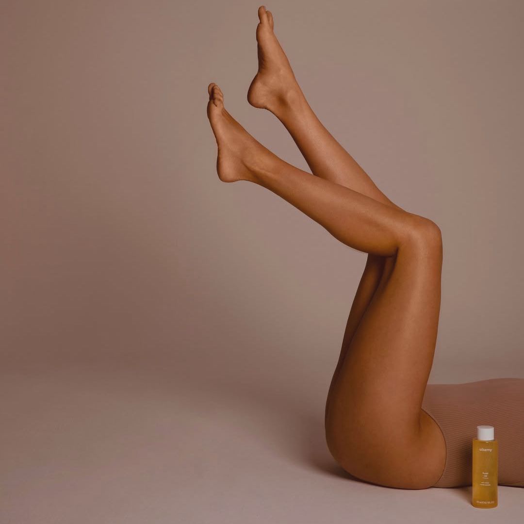 Gambe di una persona con un flacone di olio Alkemy Karité accanto.