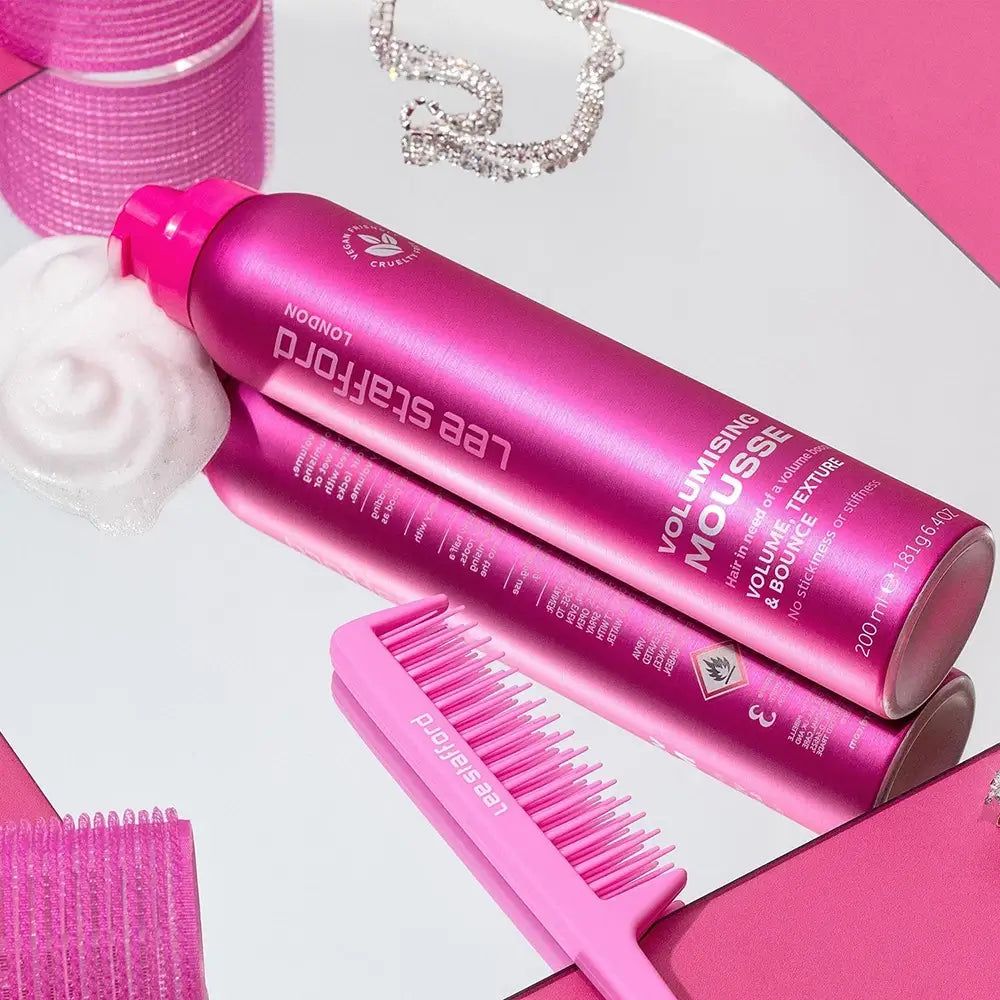 Latta rosa di Lee Stafford Volumising Mousse su specchio, con pettine e gioielli accanto. Schiuma accanto.