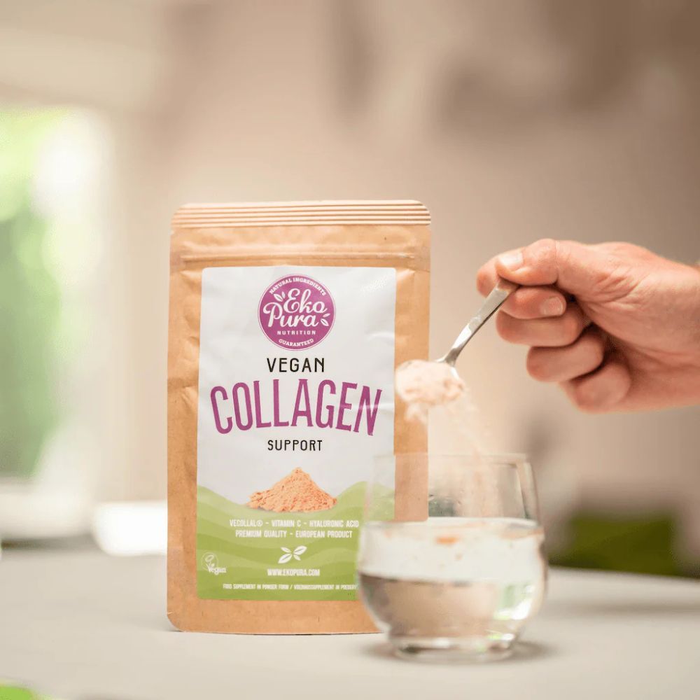 Busta Vegan Collagen Support. Cucchiaio con polvere sopra il bicchiere. Mano che tiene il cucchiaio. Sfondo: cucina.