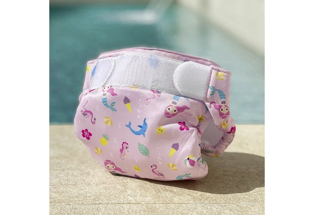 Pannolino da bagno rosa con motivi di sirene ed estivi. Chiusure in velcro. Appoggiato sul bordo piscina.
