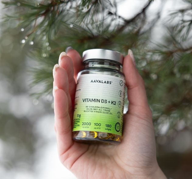 Una mano tiene una bottiglia di Aavalabs Vitamina D3+K2 Softgels davanti a uno sfondo sfocato di rami di pino.