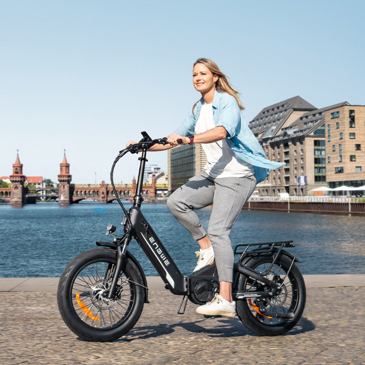 Donna in bicicletta su una e-bike ENGWE L20 3.0 Pro nera. Sfondo: acqua ed edifici.
