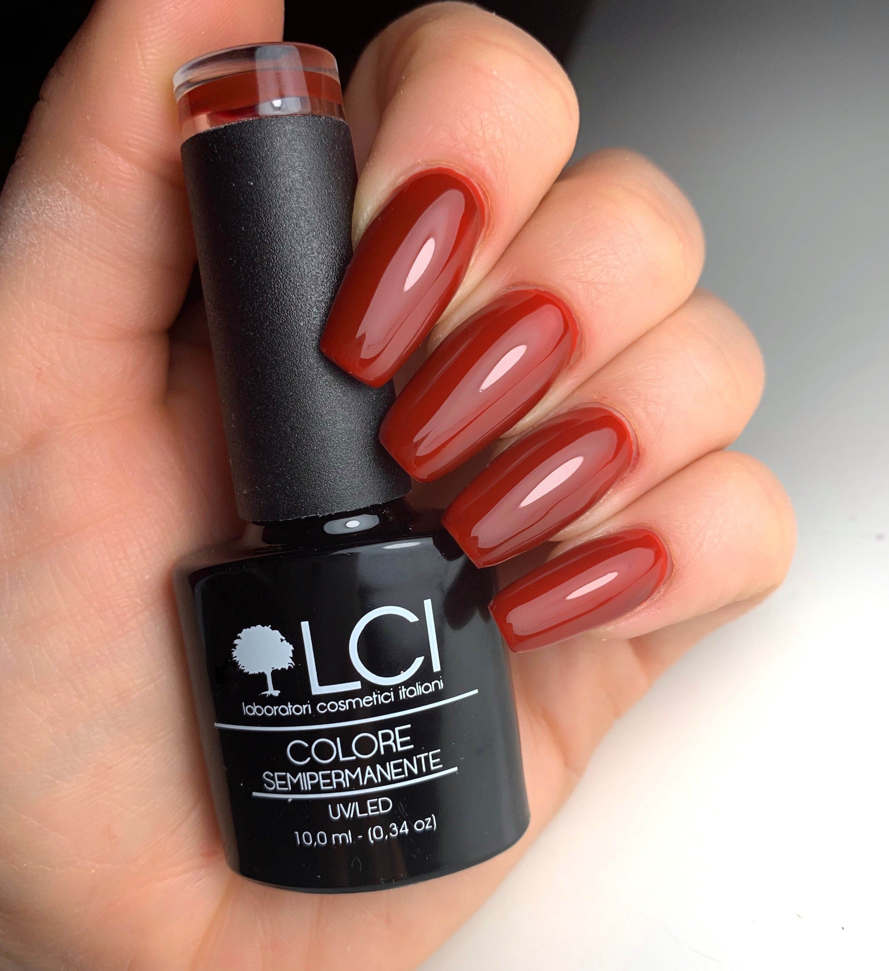 Mano con unghie smaltate e flacone di smalto. Unghie rosse e flacone di smalto nero. Scritta: LCI, Colore Semipermanente.