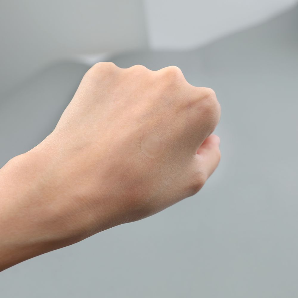 Mano chiusa con un piccolo cerotto rotondo sul dorso. Lo sfondo è grigio.