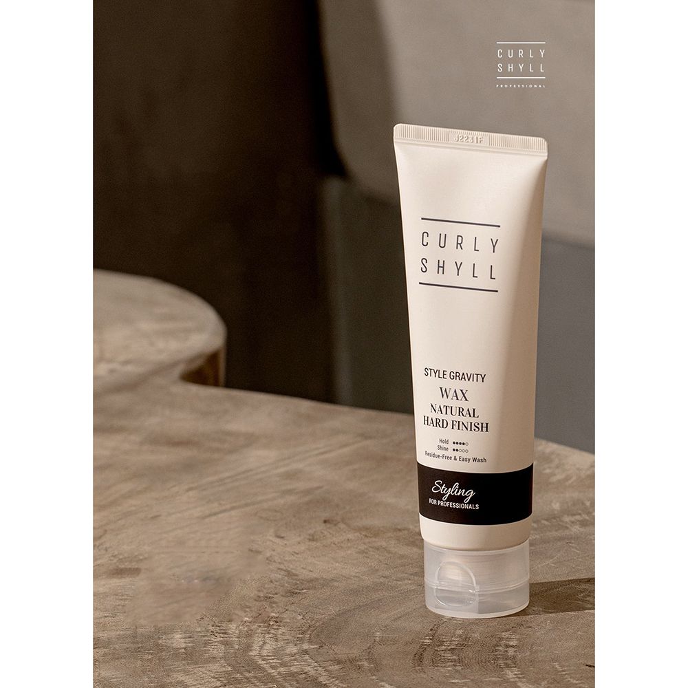 Tubo beige con testo nero. Contiene "CURLY SHYLL", "Style Gravity Wax Natural Hard Finish". Su superficie in legno.