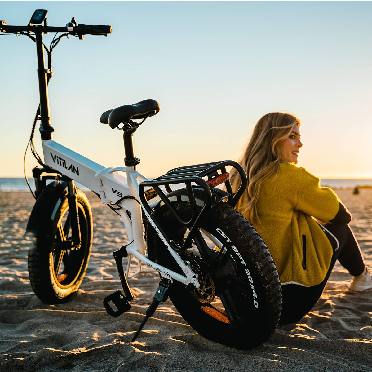 E-bike pieghevole bianca con pneumatici neri. Una donna seduta sulla spiaggia accanto alla bici. Sul telaio è scritto Vitilan V3.