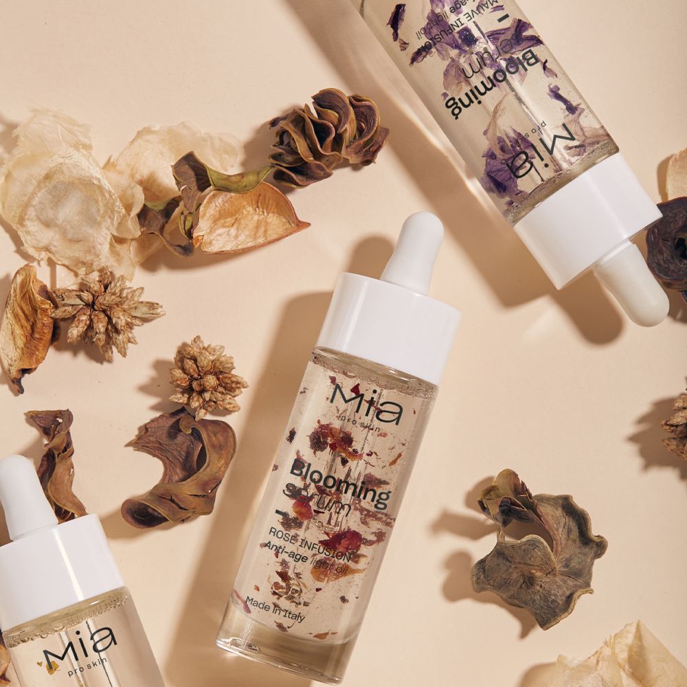 Tre flaconi di siero con contagocce. Trasparenti, con petali viola. Scritta: Mia, Blooming Serum, Mauve Infusion.