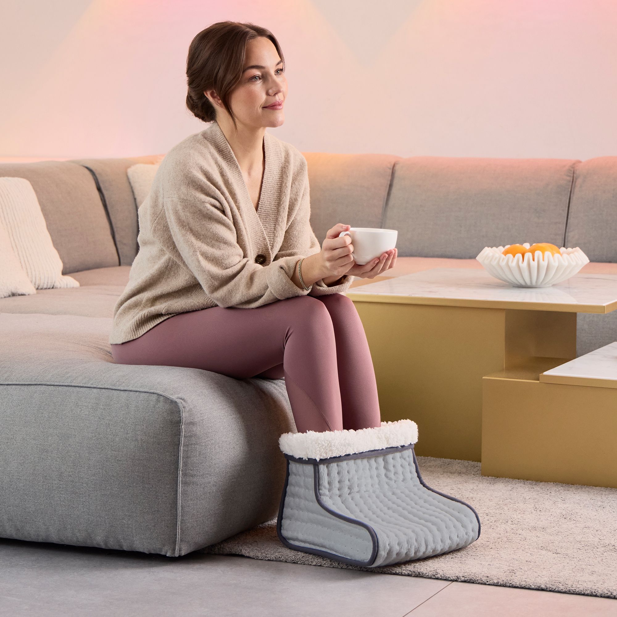 Una donna è seduta su un divano e tiene una tazza. I suoi piedi sono in uno scaldapiedi grigio con fodera interna bianca.