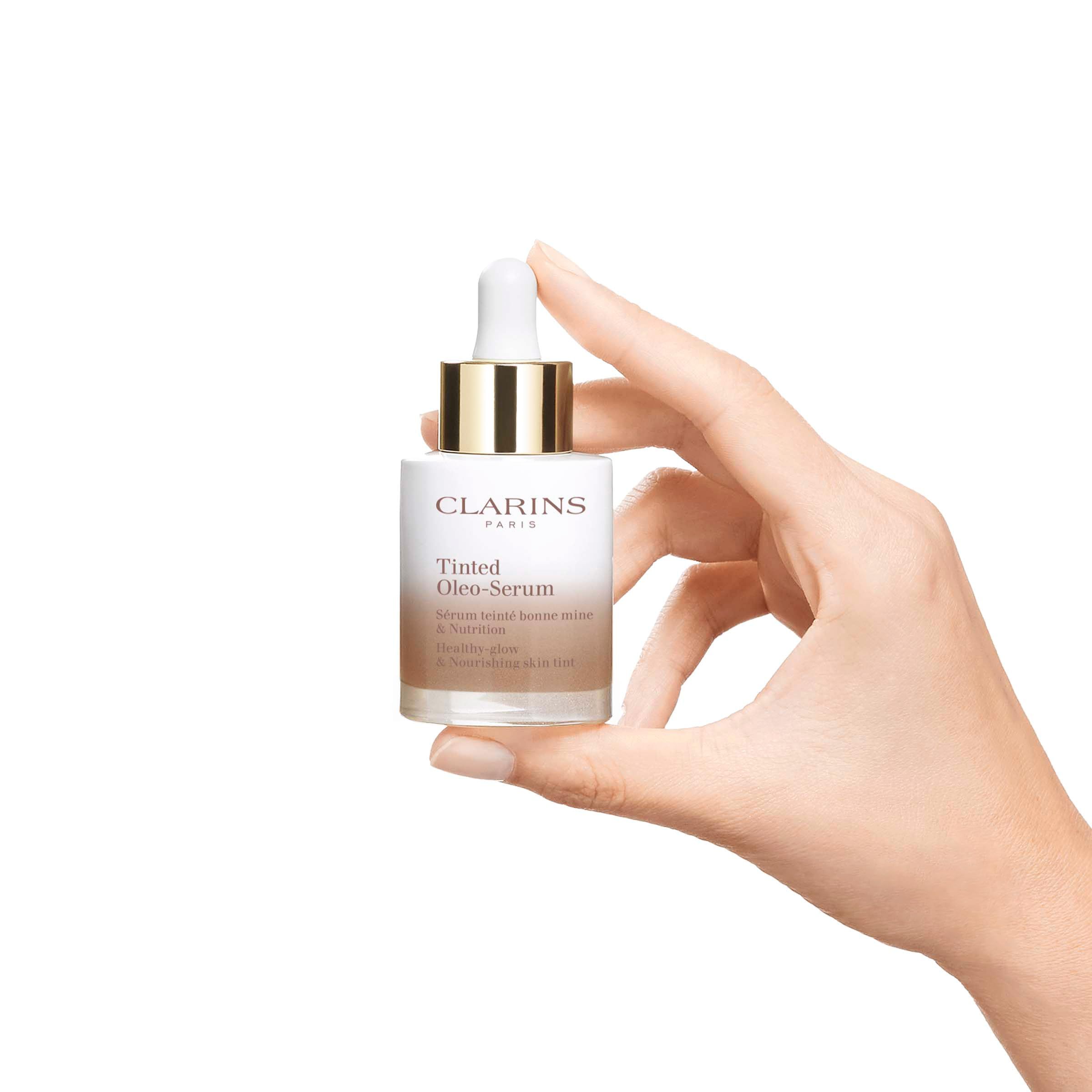 Mano che tiene un flacone Clarins Tinted Oleo-Serum. Flacone in vetro sfumato bianco-marrone, tappo dorato e contagocce bianco.