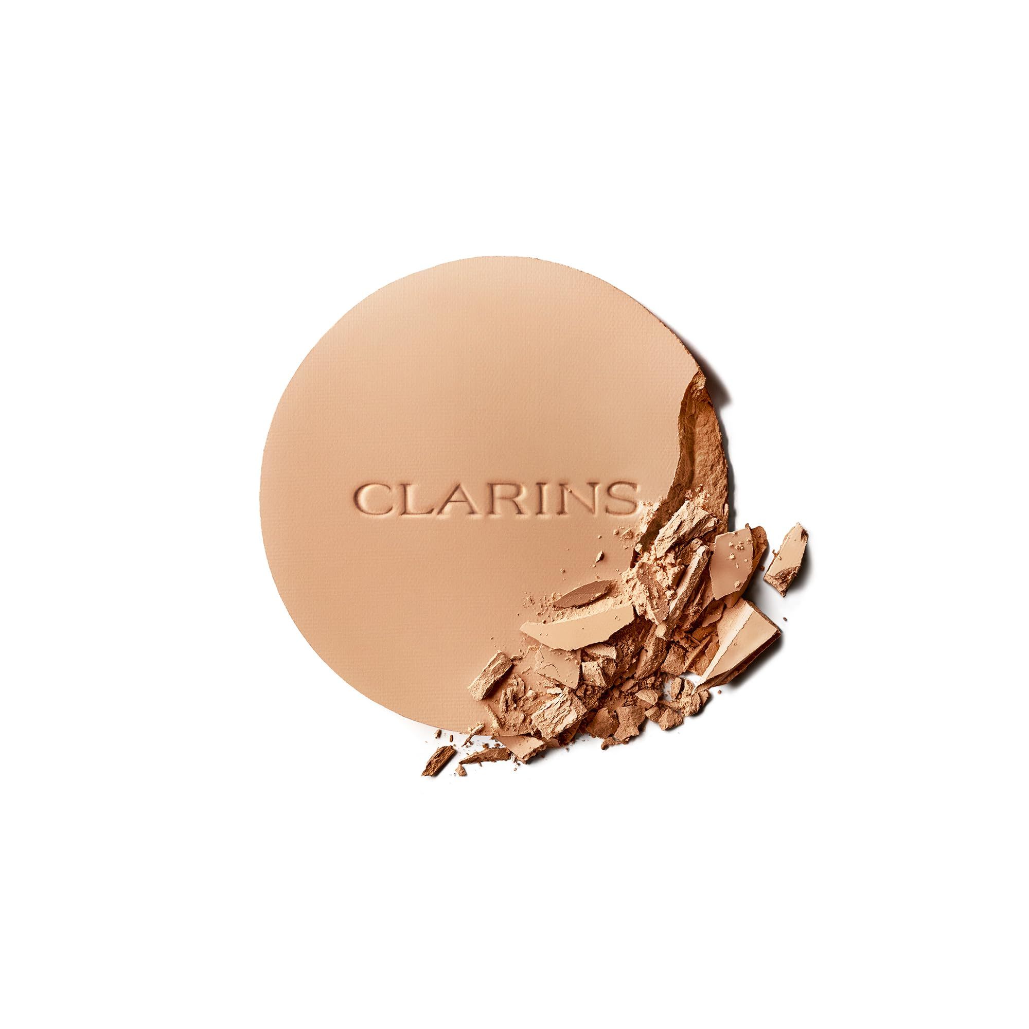 Cipria compatta con logo CLARINS. Parzialmente rotta, mostra la texture. Forma rotonda, colore chiaro.