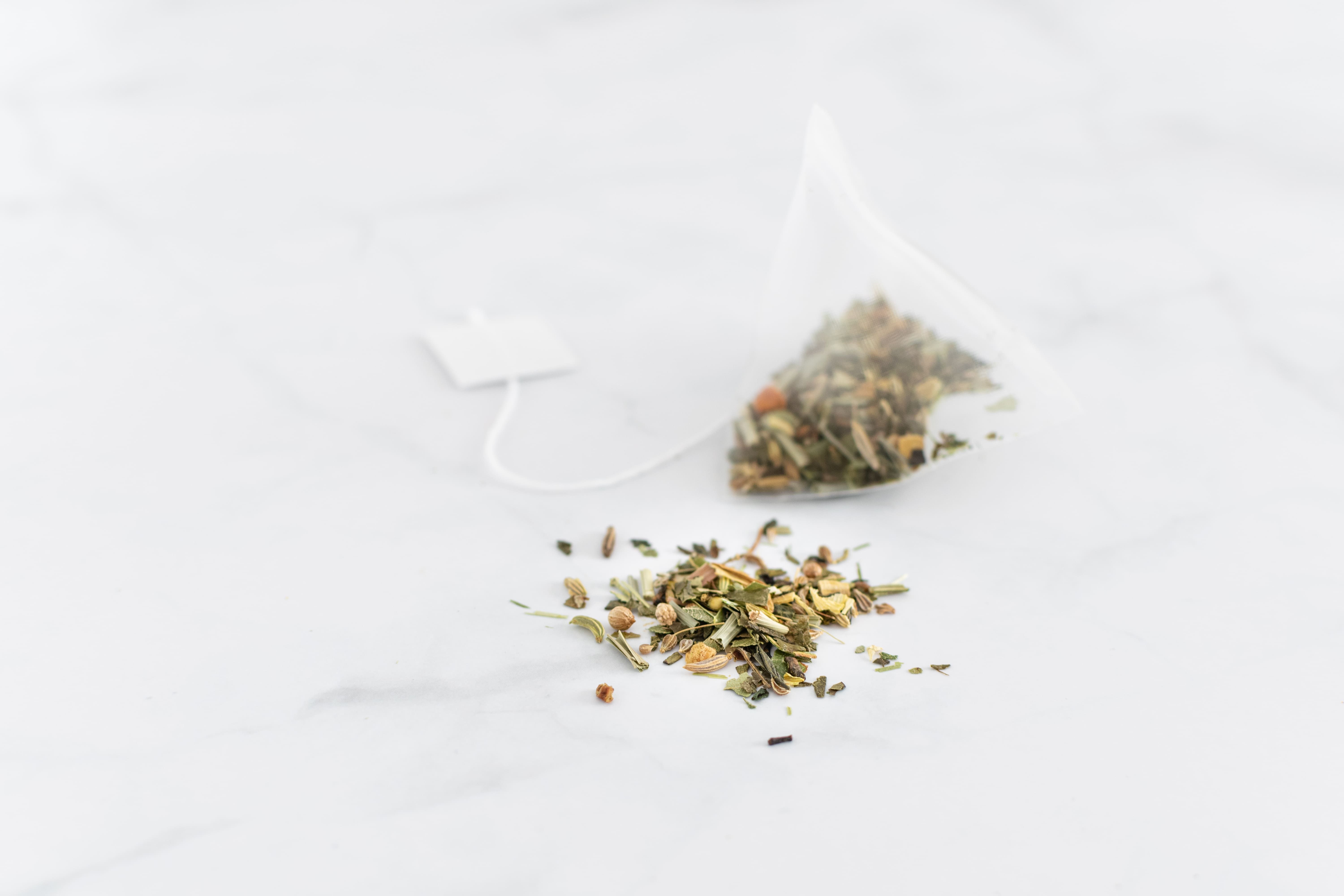 Bustina di tè piramidale con erbe. Erbe sfuse accanto. Filo bianco con etichetta.