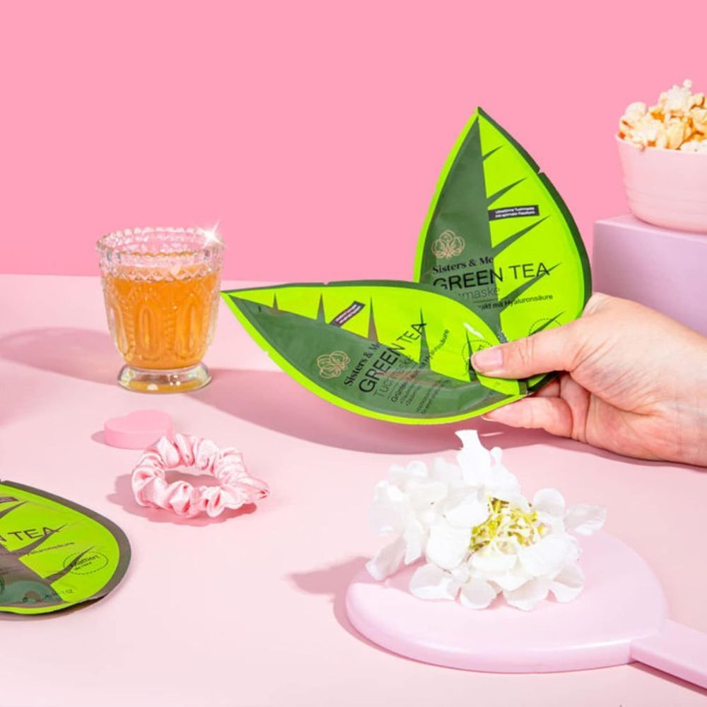 Due confezioni verdi a forma di foglia. Testo: Sisters & Me Green Tea. Su superficie rosa. Altri accessori.