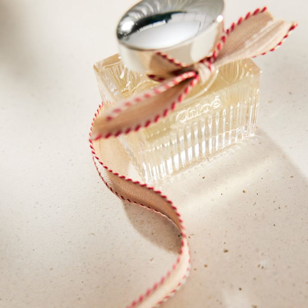 Flacone in vetro con tappo argentato e fiocco. Forma rettangolare. Il nome del marchio non è visibile.