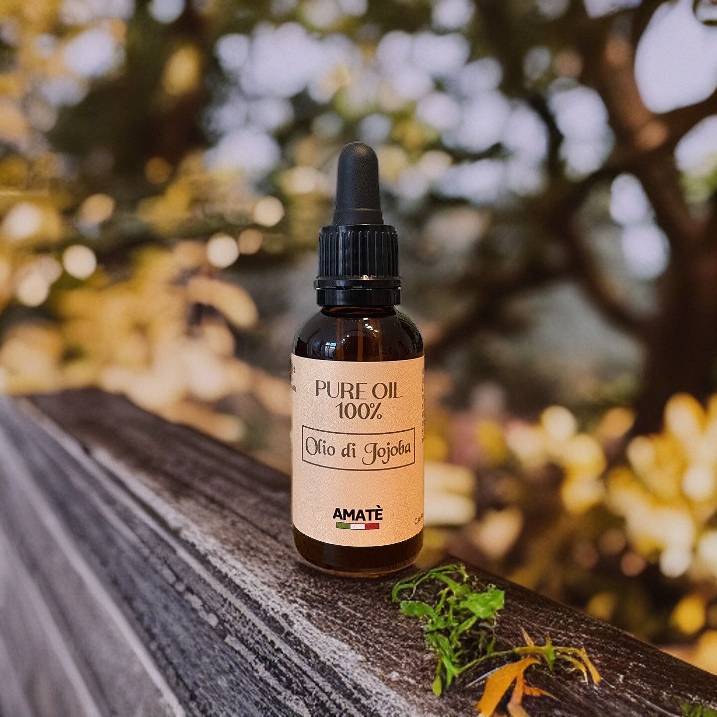 Flacone di vetro marrone con contagocce nero su legno. Etichetta "PURE OIL 100%" e "Olio di Jojoba". Marchio: AMATÈ. Sfondo: natura sfocata.