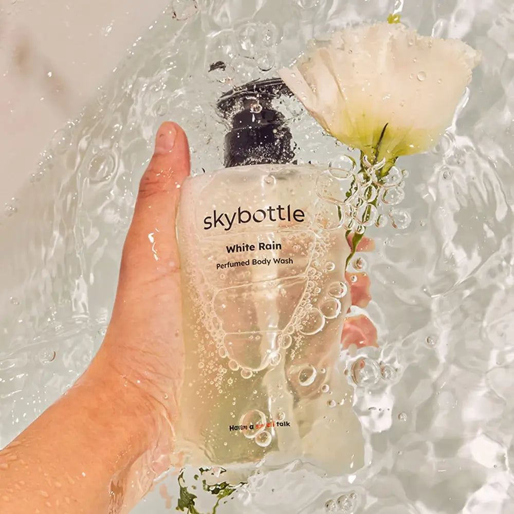 Flacone trasparente di gel doccia SKYBOTTLE White Rain con dosatore nero, immerso nell'acqua, con fiore.