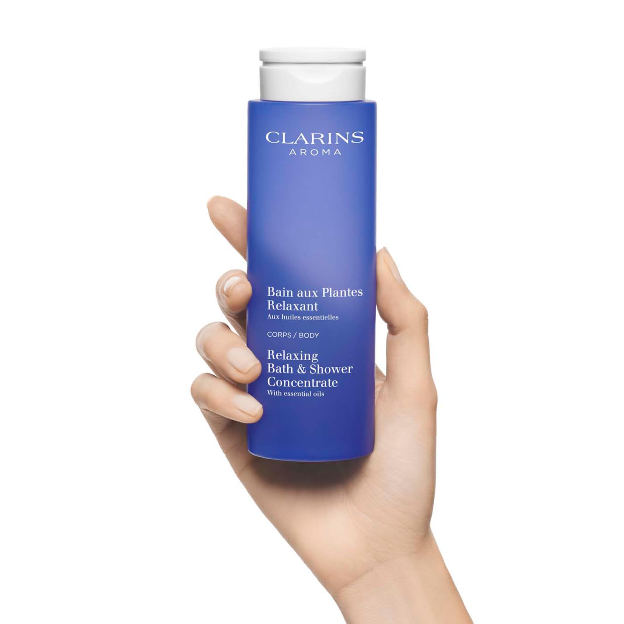 Mano che tiene un flacone blu con tappo bianco. Testo: Clarins Aroma, Bain aux Plantes Relaxant, Relaxing Bath & Shower Concentrate.
