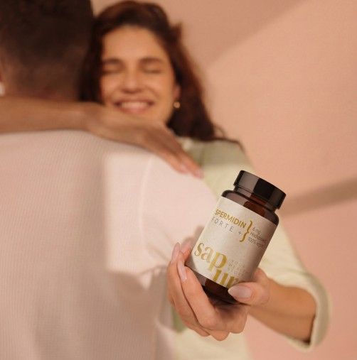 Persona che tiene in mano un flacone marrone di capsule di spermidina. Scritta: Spermidina Forte +, 100% vegano. Logo SAPURA. La persona abbraccia un'altra.