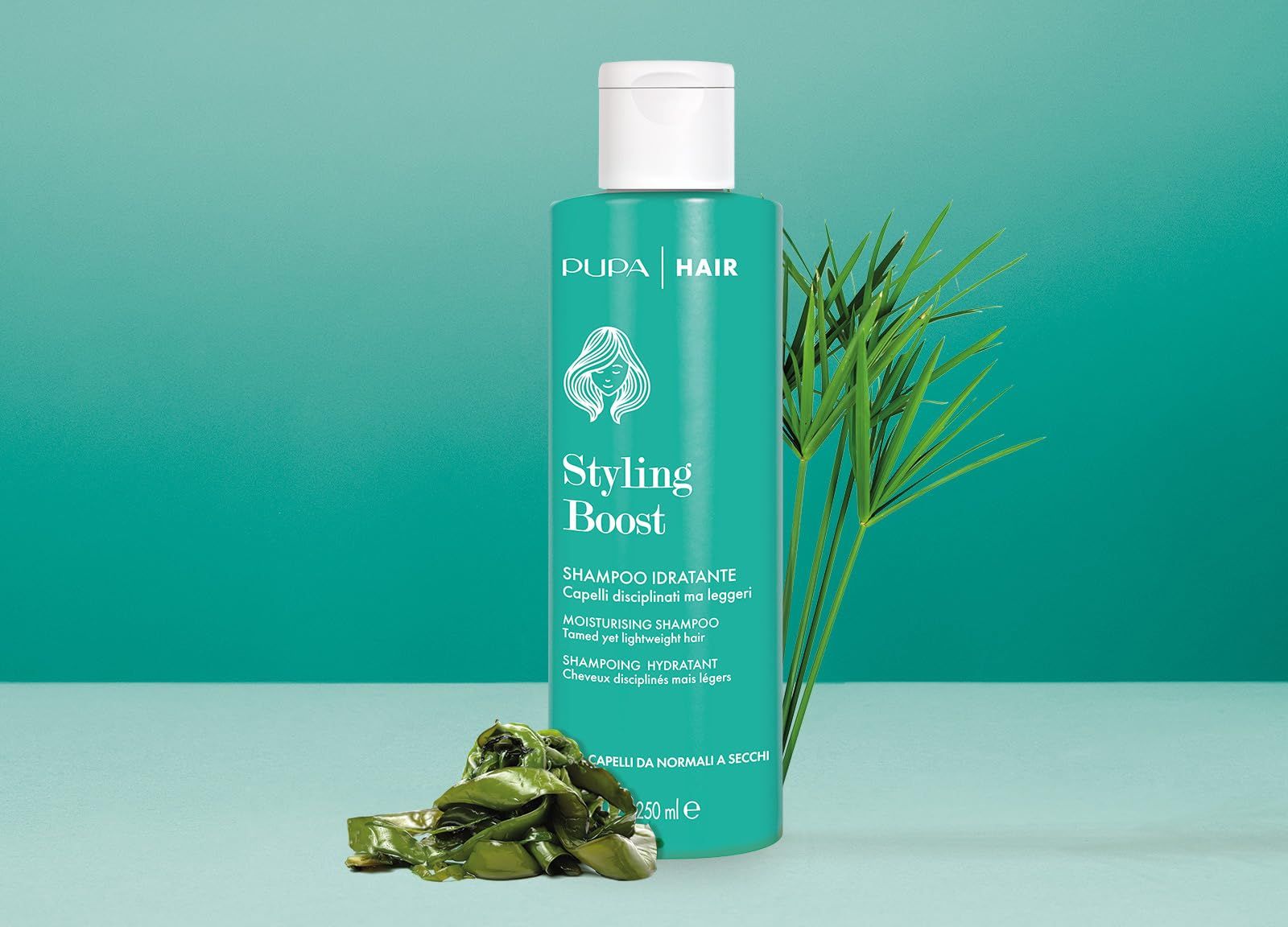 Flacone di shampoo turchese con tappo bianco. Scritta: Styling Boost, Shampoo Idratante. Piante verdi accanto.