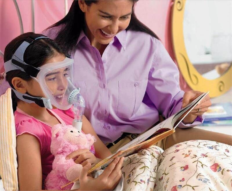 Bambino con maschera CPAP Philips, FitLife, legge a letto con un adulto.