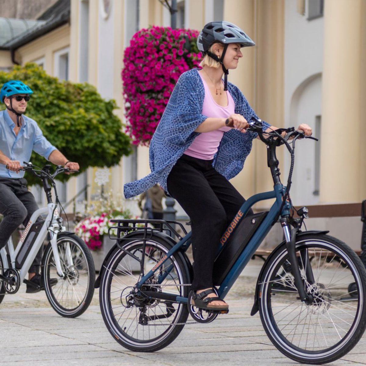 Due persone in sella a e-bike blu e bianca. Caschi, ambiente urbano.