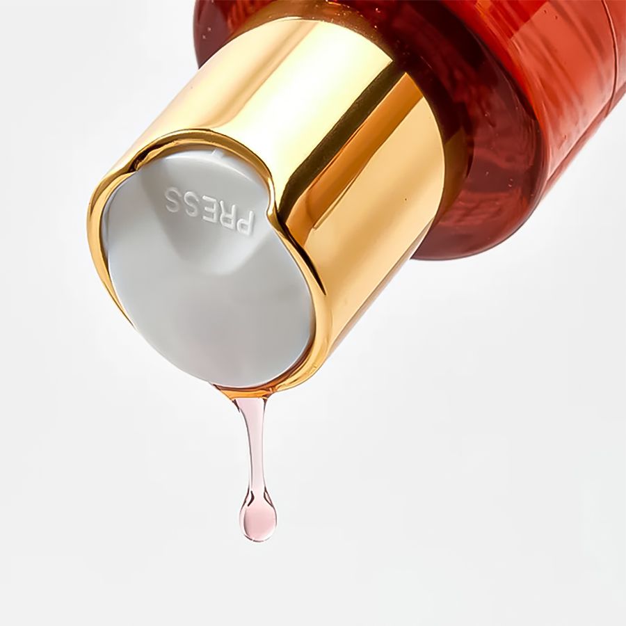 Tappo dorato con bottone grigio. Scritta: PRESS. Goccia di liquido rosa.