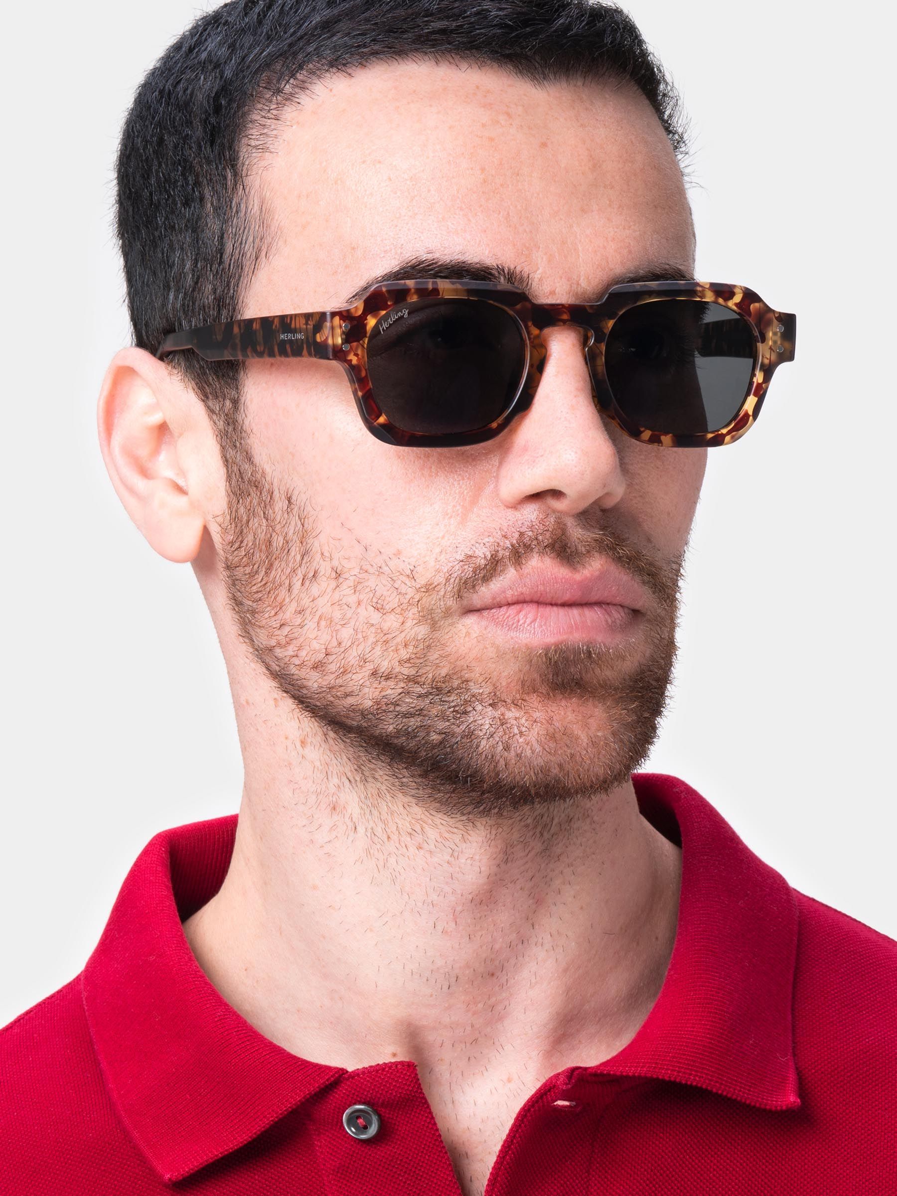Uomo con occhiali da sole marroni a fantasia tartarugata e lenti scure. Forma squadrata. Camicia rossa.