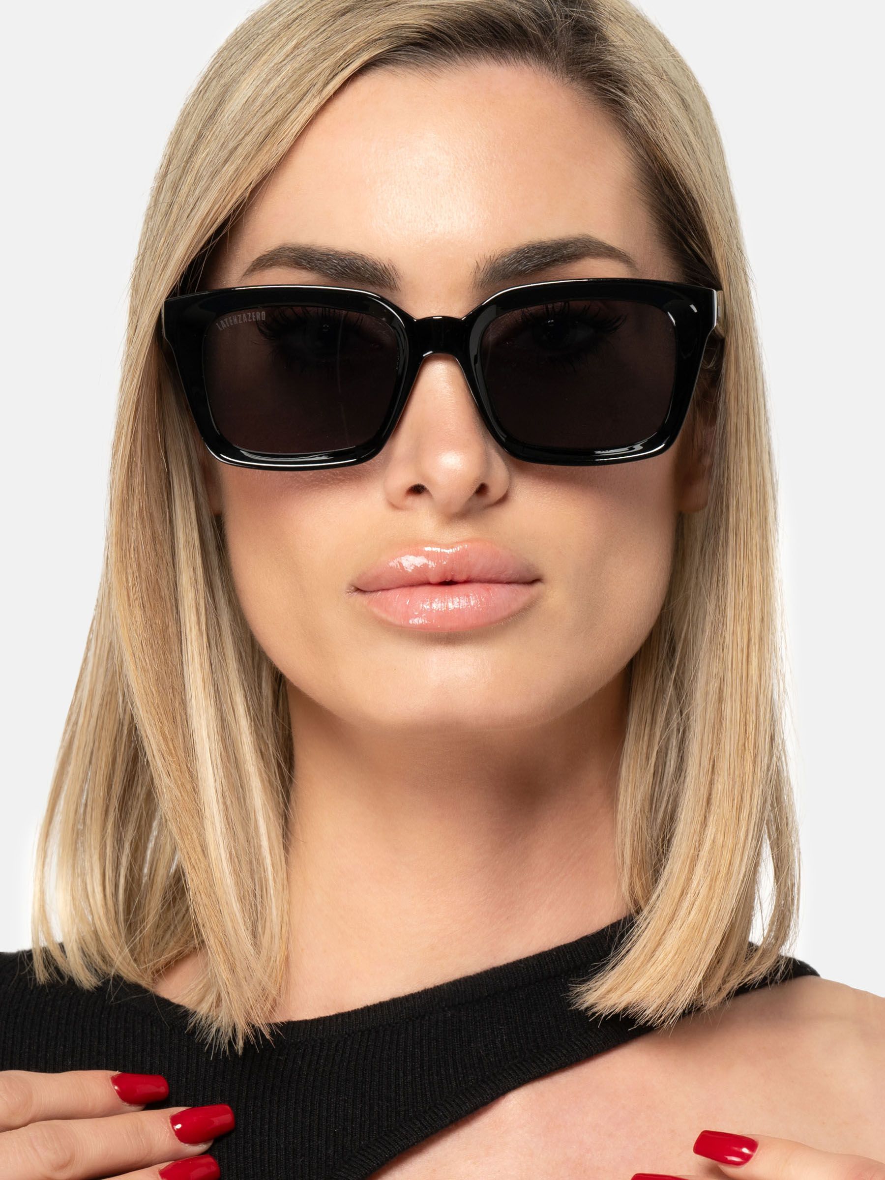 Donna con occhiali da sole neri. Lenti quadrate, montatura nera. Marchio sulla montatura. Capelli biondi.