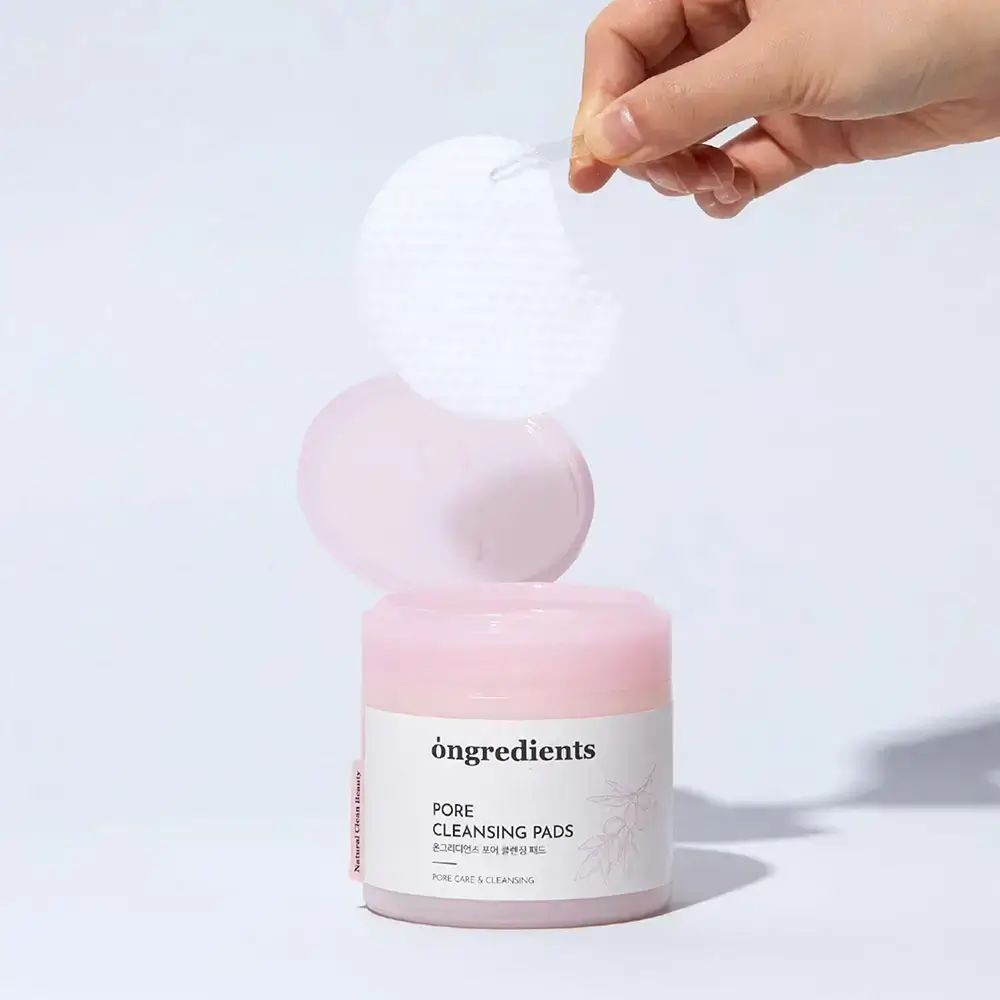 Contenitore di Pore Cleansing Pads. Coperchio rosa, etichetta bianca. Due pads tenuti da una mano. Sfondo rosa.