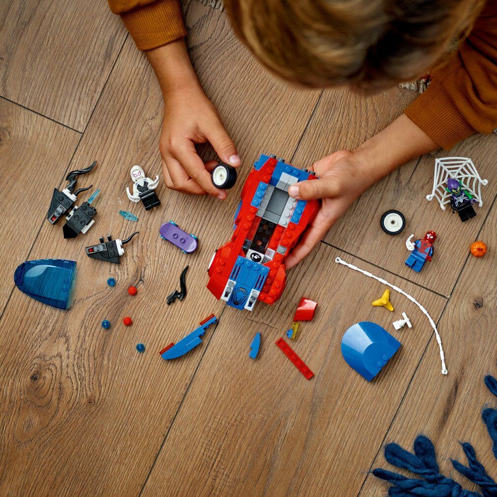 Parti LEGO Marvel smontate su pavimento in legno. Auto rossa e blu, personaggi, ruote, ragnatela e altri elementi.