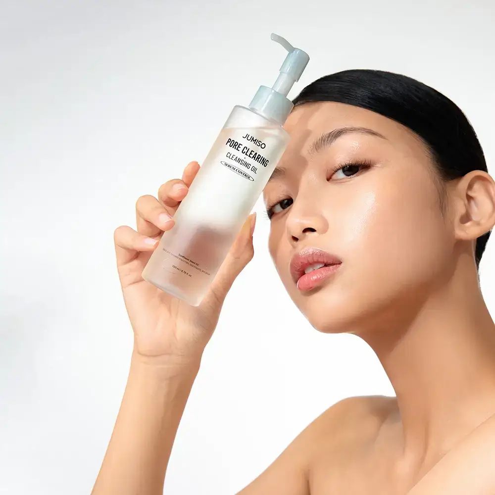 Donna tiene in mano un flacone. Scritta: JUMISO PORE CLEARING CLEANSING OIL. Flacone con dosatore.