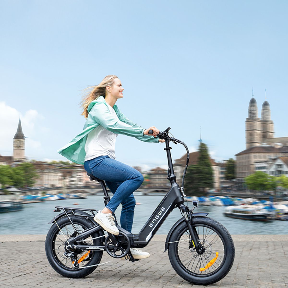 Donna in sella a una bicicletta elettrica nera. Il logo ENGWE è visibile. Sfondo: edifici e acqua.