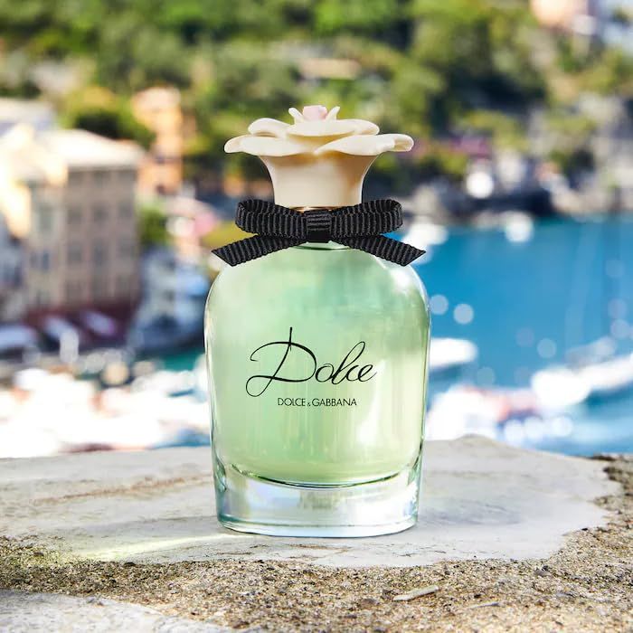 Flacone di profumo con fiore bianco, fiocco nero e scritta Dolce. Sfondo con mare ed edifici.