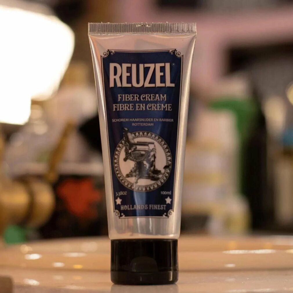 Tubo d'argento con etichetta blu. Scritta REUZEL FIBER CREAM. Immagine di una poltrona da barbiere. Tappo nero.